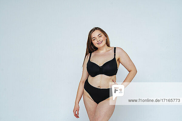 Lächelnde kurvige junge Frau trägt schwarzen Bikini posiert gegen weißen Hintergrund
