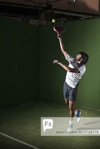 Male athlete servicing ball while playing padel tennis in sports court at night