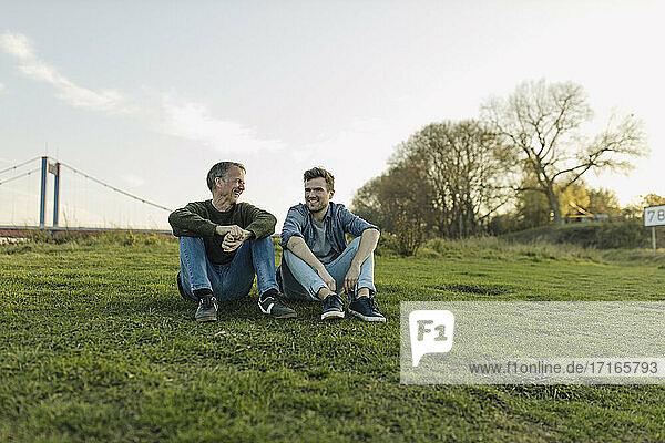 Happy father and son talking with each other while sitting on grass against sky