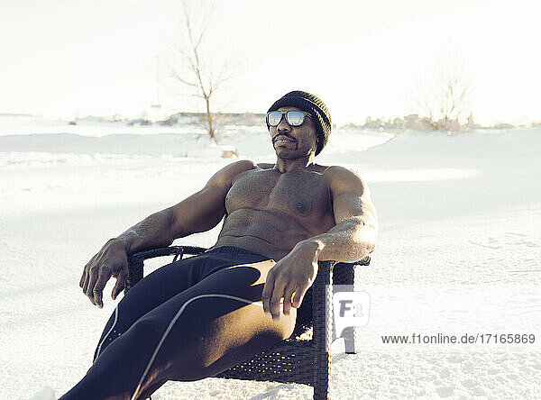 Männlicher Sportler ohne Hemd schaut weg  während er sich auf einem Stuhl im Schnee entspannt