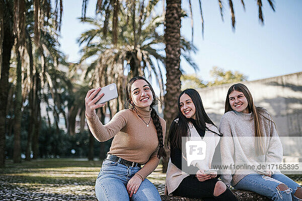 Lächelndes Teenager-Mädchen  das ein Selfie mit Freunden macht  während es gegen Bäume im Park sitzt