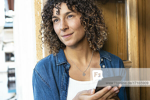 Curly woman with smart phone looking away at home