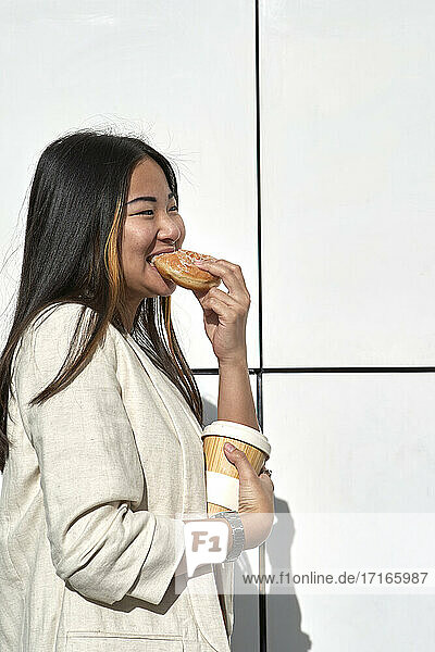 Smiling young woman with coffee cup eating doughnut against wall during sunny day