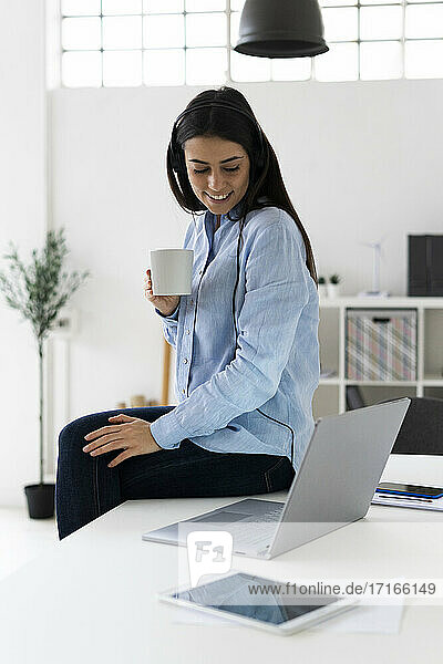 Unternehmerin mit Mikrofon-Headset  die Kaffee trinkt  während sie über ihren Laptop im Büro eine Telefonkonferenz führt