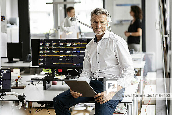Männlicher Unternehmer mit digitalem Tablet auf dem Schreibtisch sitzend mit Kollegen im Hintergrund in einem Großraumbüro