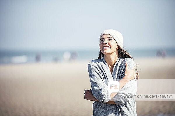 Lächelnde Frau  die sich selbst umarmt  während sie am Strand an einem sonnigen Tag steht