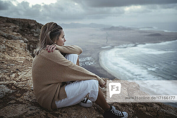 Junge Frau schaut auf den Strand von Famara  während sie auf einem Berg in Lanzarote  Spanien  sitzt