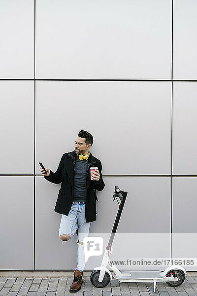 Handsome man using smart phone while standing by electric push scooter against wall