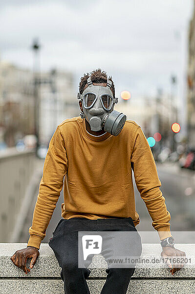Young man wearing gray gas mask sitting on retaining wall