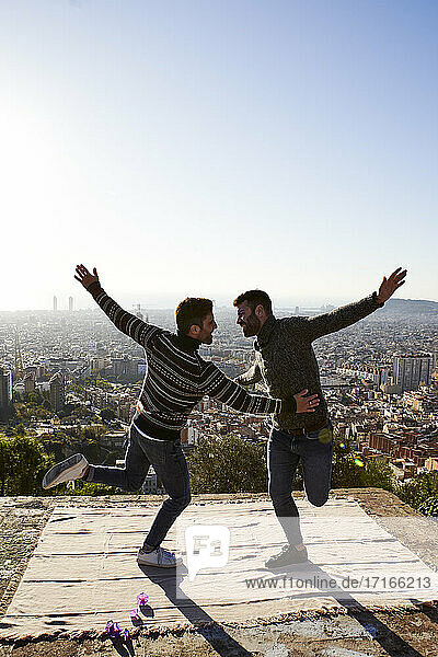 Cheerful gay couple with arms outstretched dancing on observation point  Bunkers del Carmel  Barcelona  Spain