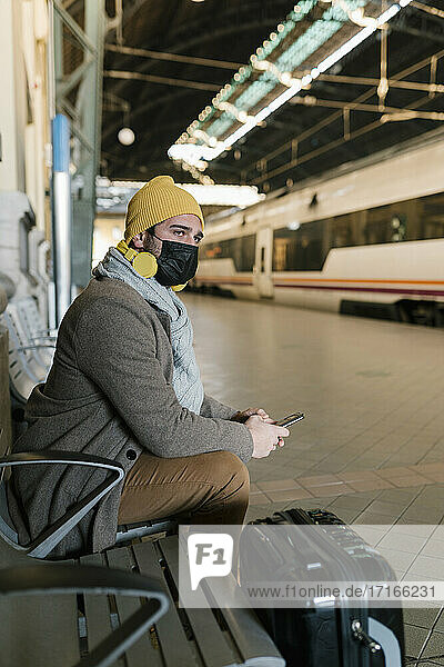 Mann mit Gesichtsmaske und Mobiltelefon  der auf einer Bank am Bahnhof sitzt und wartet