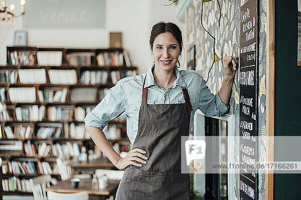 Smiling female owner with hand on hip leaning by wall at coffee shop