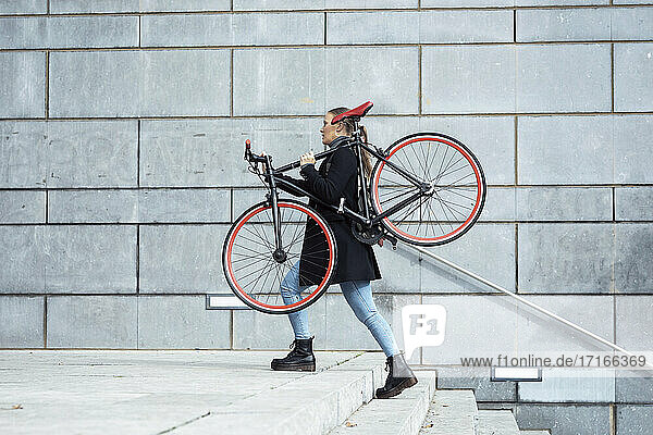 Woman carrying cycle while walking on staircase by wall