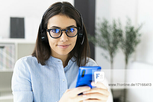 Lächelnde Geschäftsfrau mit Brille und Mikrofon-Headset  die ein Mobiltelefon benutzt  während sie im Büro sitzt