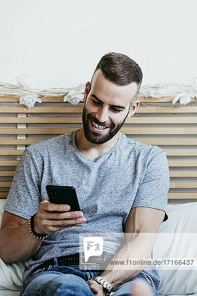 Lächelnder junger Mann  der zu Hause auf dem Bett sitzend an seinem Mobiltelefon arbeitet
