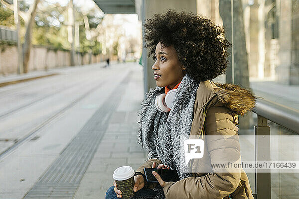 Afro-Frau mit Smartphone und Einweg-Kaffeebecher nachdenklich am Busbahnhof im Winter