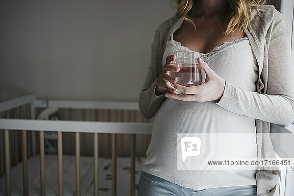 Nahaufnahme einer schwangeren Frau  die ein Trinkglas hält  während sie sich zu Hause an ein Kinderbett lehnt