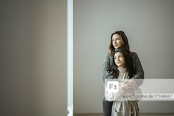 Mother and daughter looking away while embracing each other against wall at home