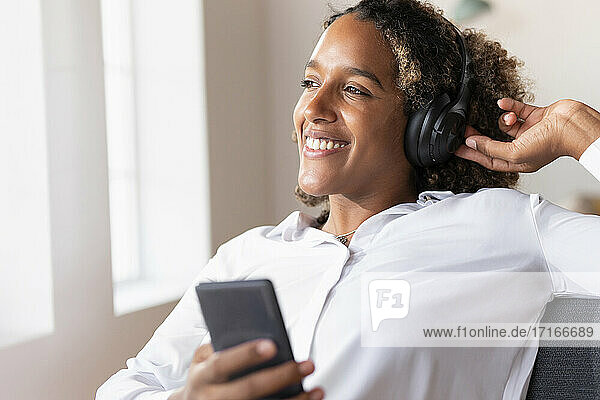 Glückliche Frau mit Kopfhörern  die ein Mobiltelefon benutzt  während sie zu Hause sitzt