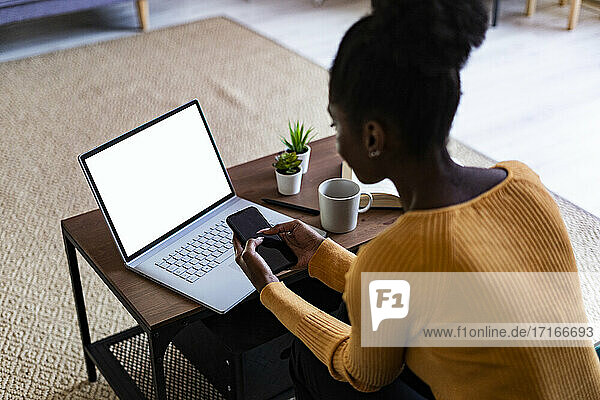 Afro woman using smart phone while sitting with laptop at home