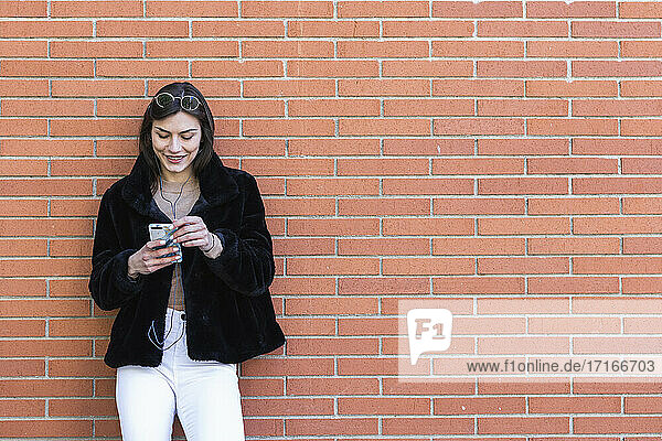 Smiling woman with mobile phone listening music while standing against brick wall