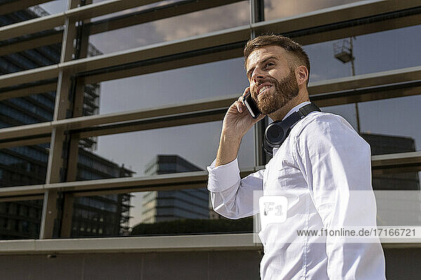 Smiling businessman talking on mobile phone while standing against building