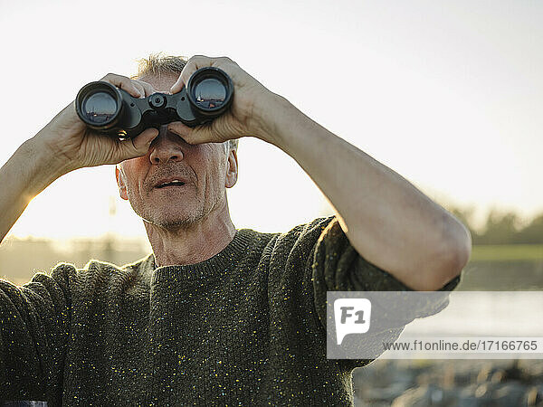 Senior man looking through binoculars against clear sky at riverbank