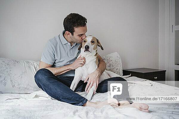 Mid adult man kissing dog while sitting on bed against wall at home