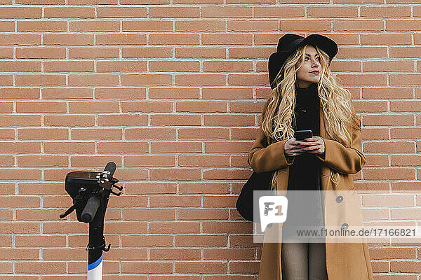 Woman in coat using smart phone while standing against brick wall