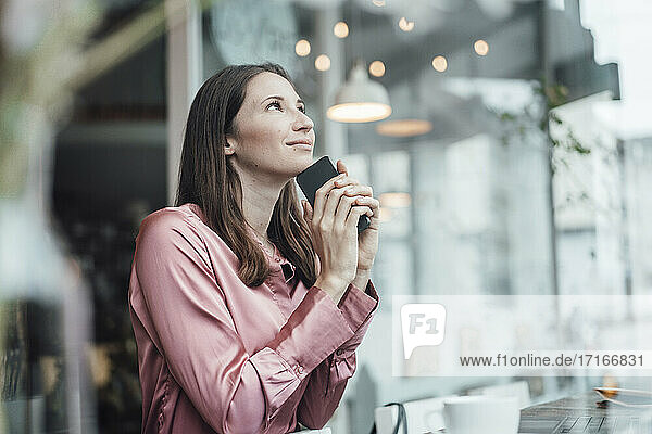 Lächelnde weibliche Fachkraft mit Mobiltelefon träumt im Cafe