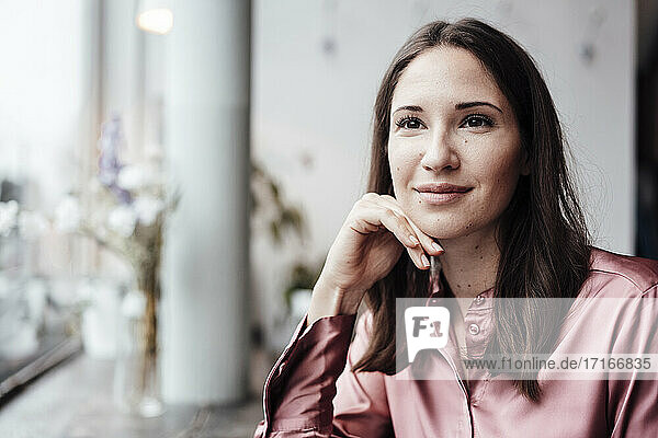Lächelnde Geschäftsfrau mit Hand am Kinn  die in einem Cafe nachdenkt