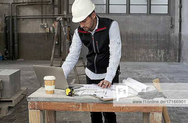 Männlicher Architekt mit Bauplänen  der einen Laptop auf einem Tisch benutzt  während er in einem Gebäude steht