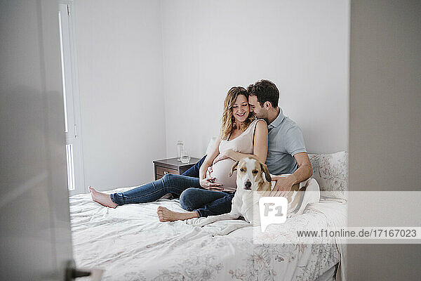 Man romancing with pregnant wife while sitting with dog on bed seen through doorway