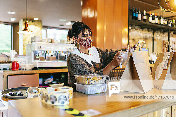 Weiblicher Besitzer in Maske  der eine Quittung auf eine braune Papiertüte auf dem Tresen eines Restaurants klebt