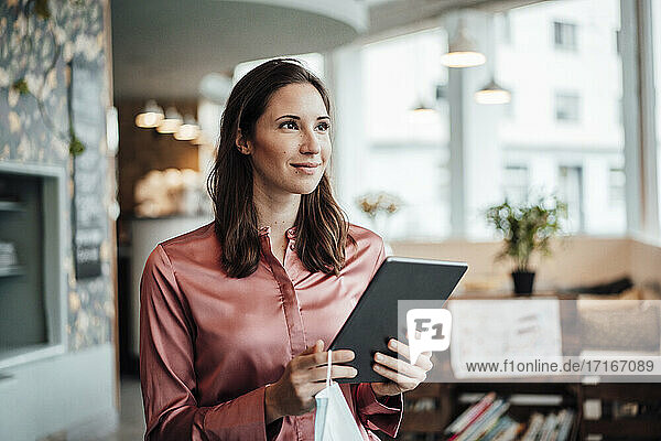 Lächelnde Unternehmerin mit digitalem Tablet  die in einem Café während einer Pandemie wegschaut