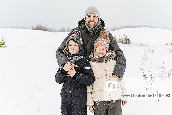 Smiling father with children standing on snow covered landscape against sky
