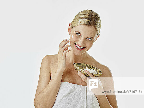 Smiling woman applying face cream while standing against white background