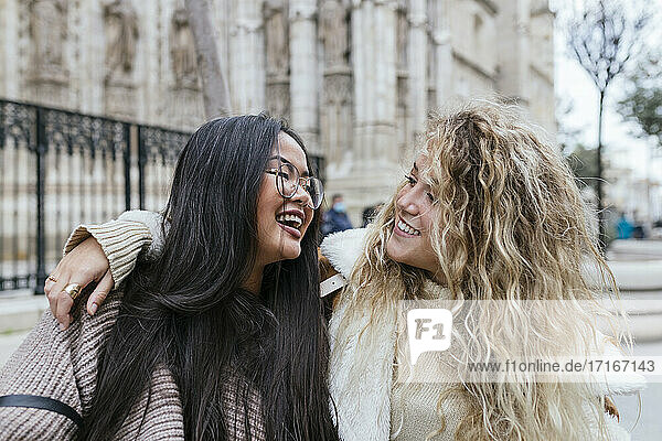 Young woman with arms around female friend smiling at each other while standing outdoors