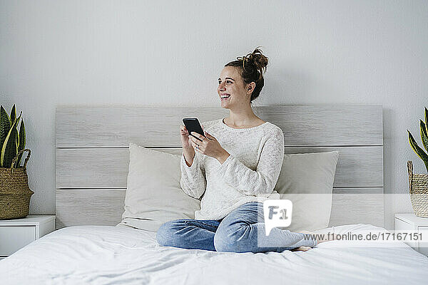 Smiling woman using mobile phone while sitting on bed at home