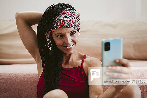 Smiling woman wearing headscarf taking selfie while sitting by sofa at home