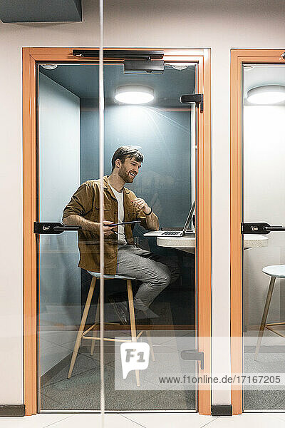 Männlicher Berufstätiger  der ein Videogespräch über einen Laptop führt  gesehen durch eine Glastür im Büro