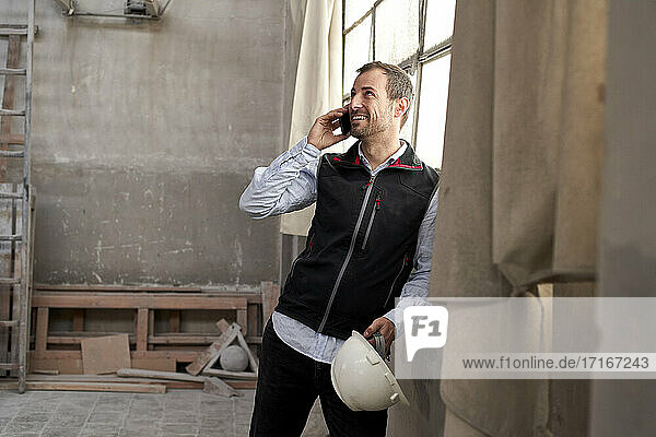 Lächelnder männlicher Bauunternehmer  der über sein Smartphone spricht  während er in einem Gebäude steht