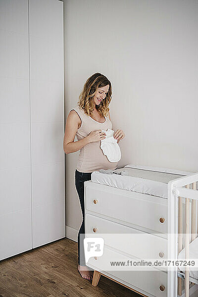 Lächelnde schwangere Frau  die Babykleidung hält  während sie zu Hause am Schrank steht