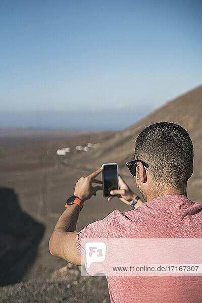 Junger männlicher Tourist fotografiert mit seinem Smartphone am Aussichtspunkt von Femés  Lanzarote an einem sonnigen Tag  Spanien