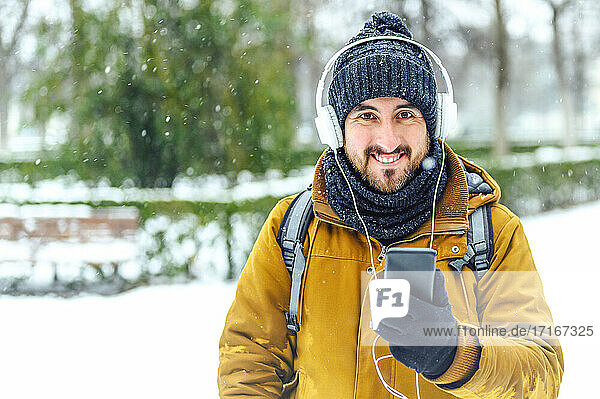 Smiling man holding mobile phone while listening music through headphones during winter