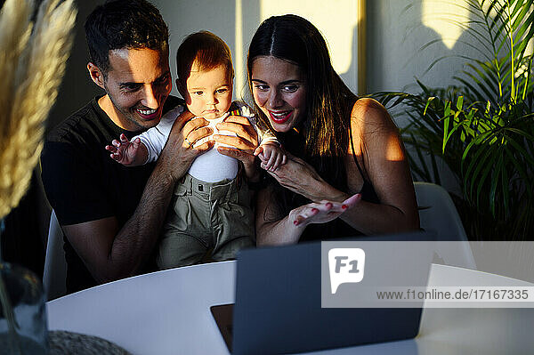 Smiling couple holding son while talking on video call through laptop sitting at home