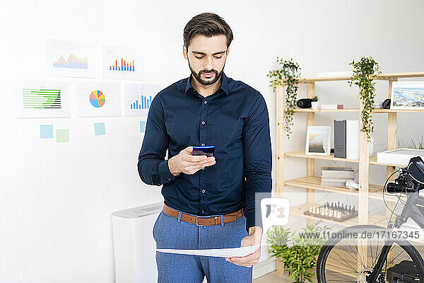 Businessman photographing document through smart phone while standing against wall in office