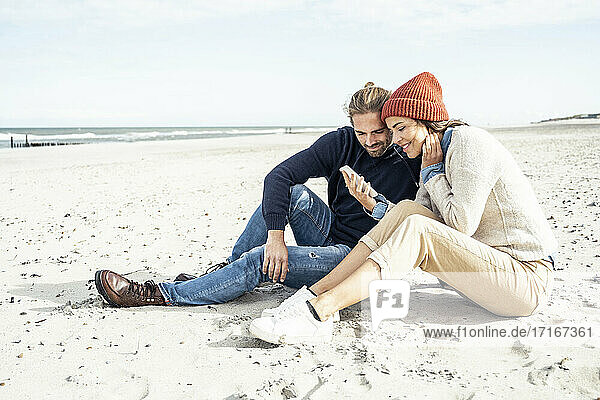 Young couple sitting together on beach sand and using smart phone