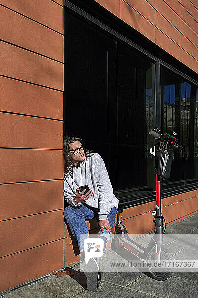 Young disabled man sitting on window sill by electric push scooter during sunny day