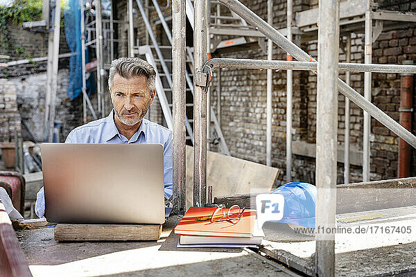 Älterer Ingenieur  der auf der Baustelle sitzend am Laptop arbeitet